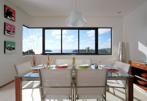Un coin repas avec une table et des chaises en verre, une grande fenêtre offrant une belle vue et des œuvres d’art accrochées au mur.