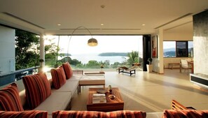 A spacious living room with a large window overlooking a body of water, a sofa set, a coffee table, and a dining area. - The Heights Penthouse 3bedroom A2 near Kata Beach (Karon)