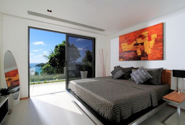 Une chambre moderne avec un grand lit, une vue sur l’extérieur grâce à une porte-fenêtre coulissante et un tableau accroché au mur.