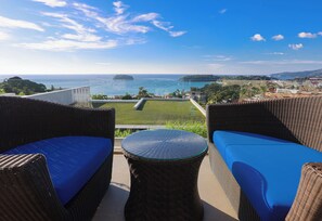 Ein Balkon mit Korbmöbeln und einem Glastisch mit Blick auf eine Küstenlandschaft mit Gebäuden und viel Grün.