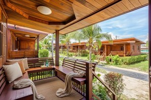 Una terraza de madera con un banco, una zona cubierta y vistas a un complejo turístico con palmeras y edificios.