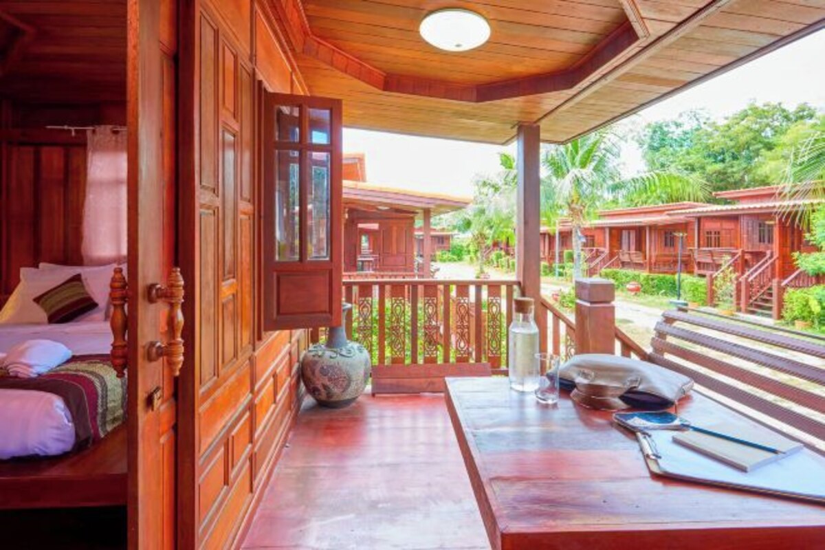 A wooden room with a bed, a table with a vase, and a view of a wooden structure outside.