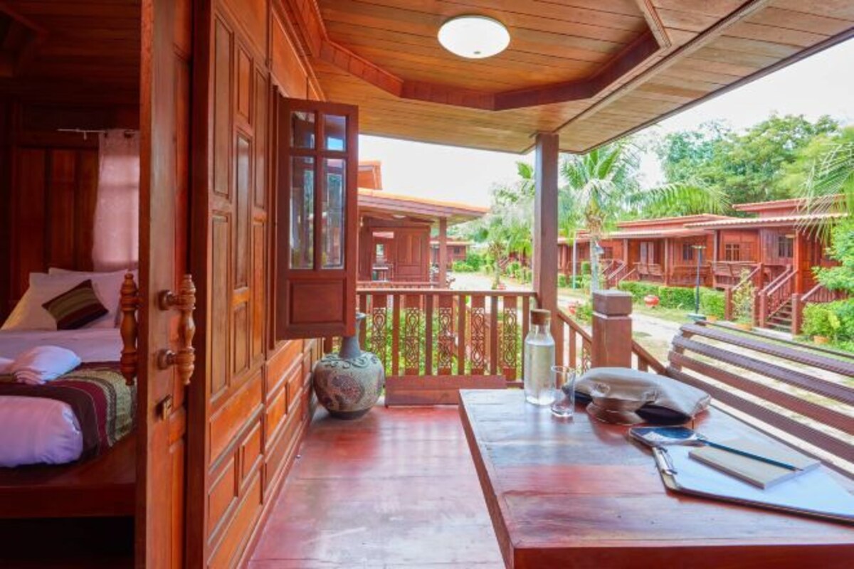 A wooden room with a bed, a table with a vase, and a view of other wooden structures and greenery.