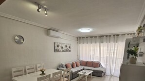 A living room with a sofa, a dining table with chairs, a coffee table, and a large plant.