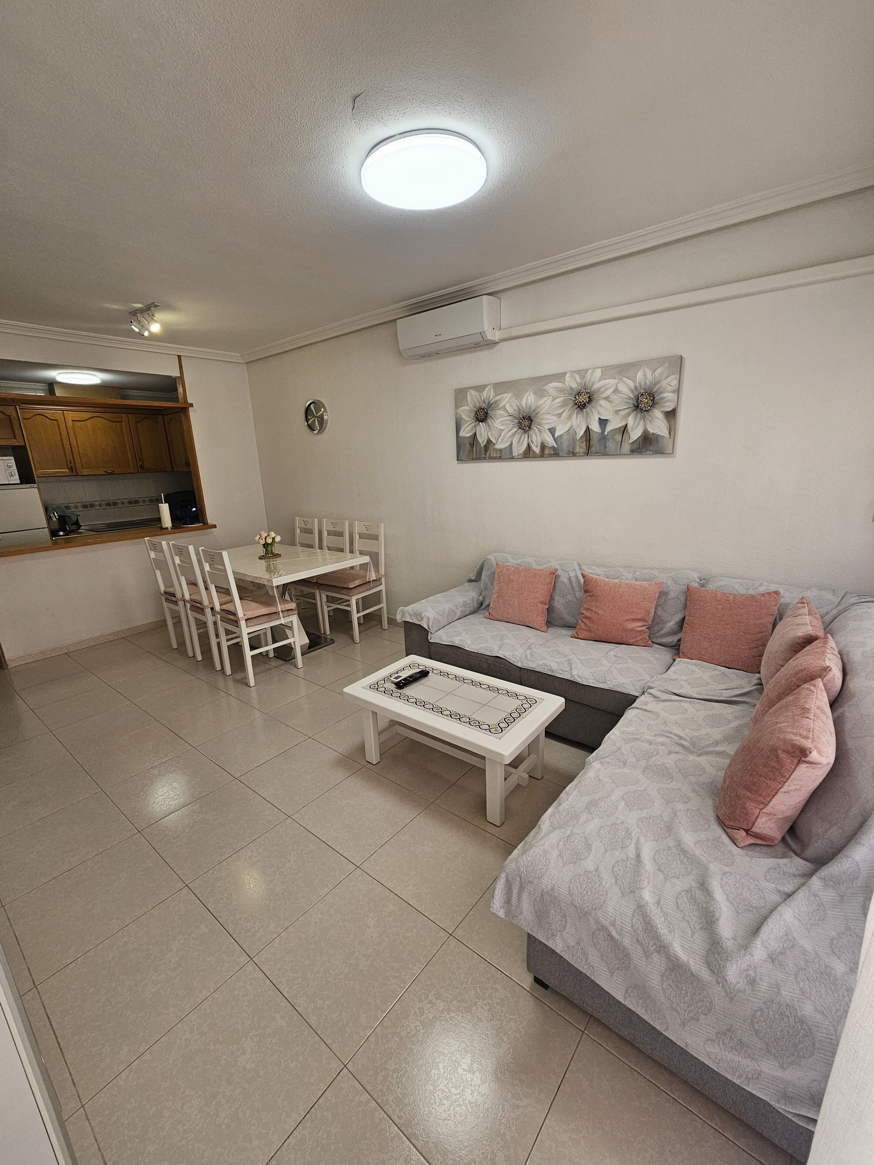 Una sala de estar con sofá, mesa de centro, mesa de comedor, sillas y una cocina con armarios y aire acondicionado.