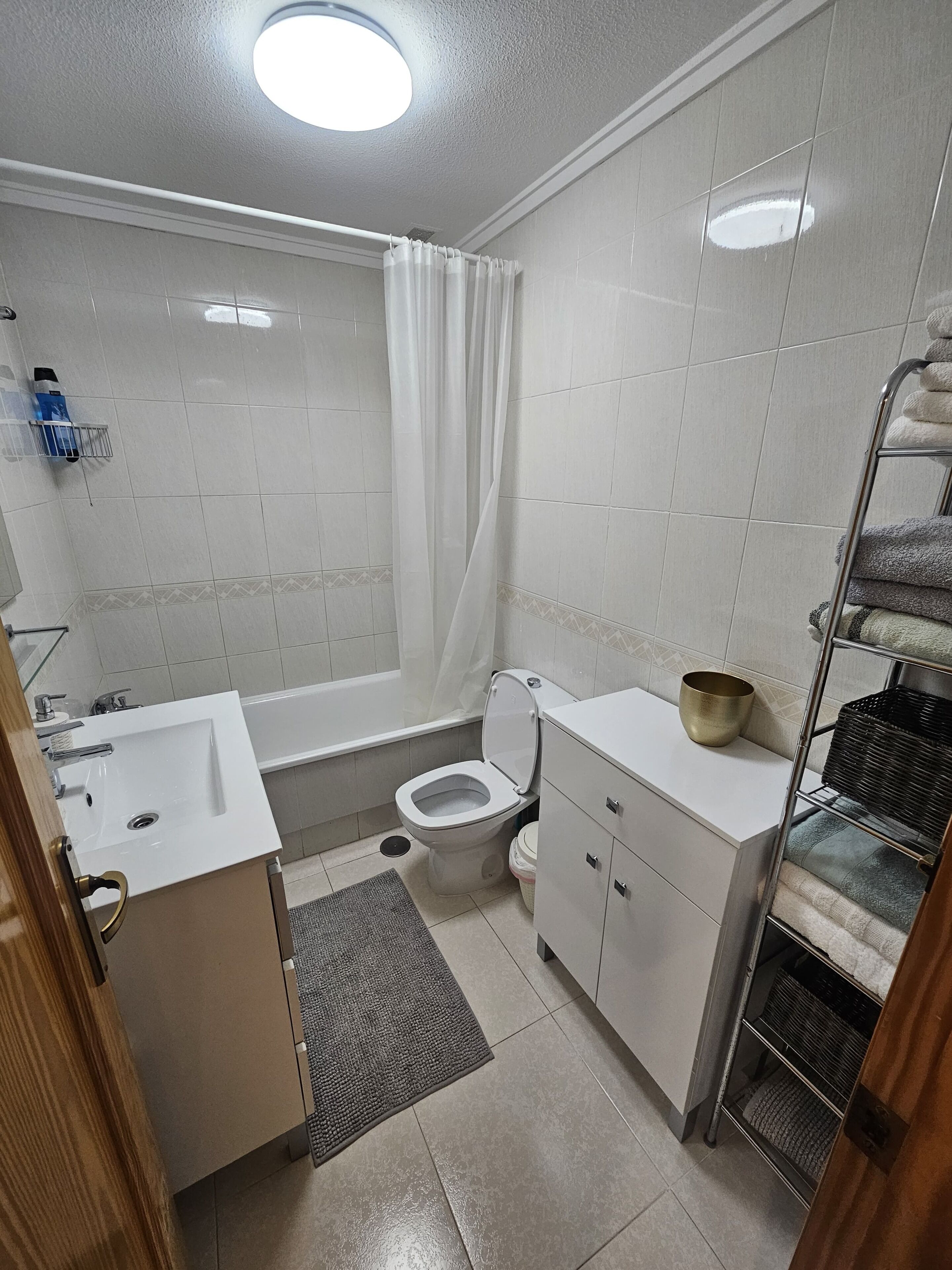 Et badeværelse med en hvid vask, toilet og badekar. Der er et bruseforhæng, et håndklædestativ og en lille hylde med en skål.