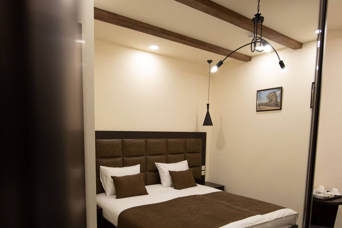 A neatly made bed with a brown headboard and white bedding, a bedside table with a lamp, and a wooden floor with a herringbone pattern.