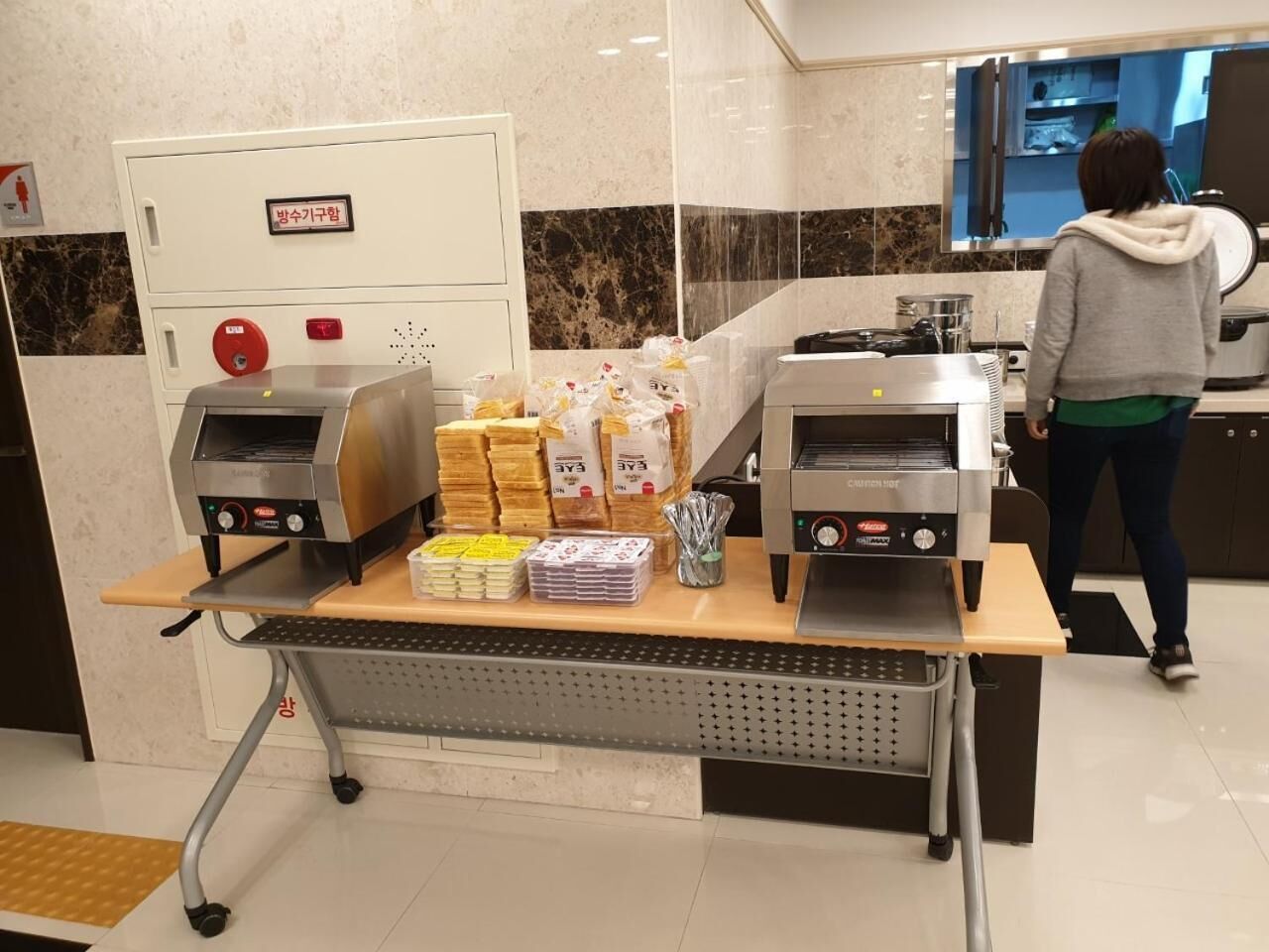 Een buffet met twee ovens, diverse verpakte maaltijden en een persoon op de achtergrond.