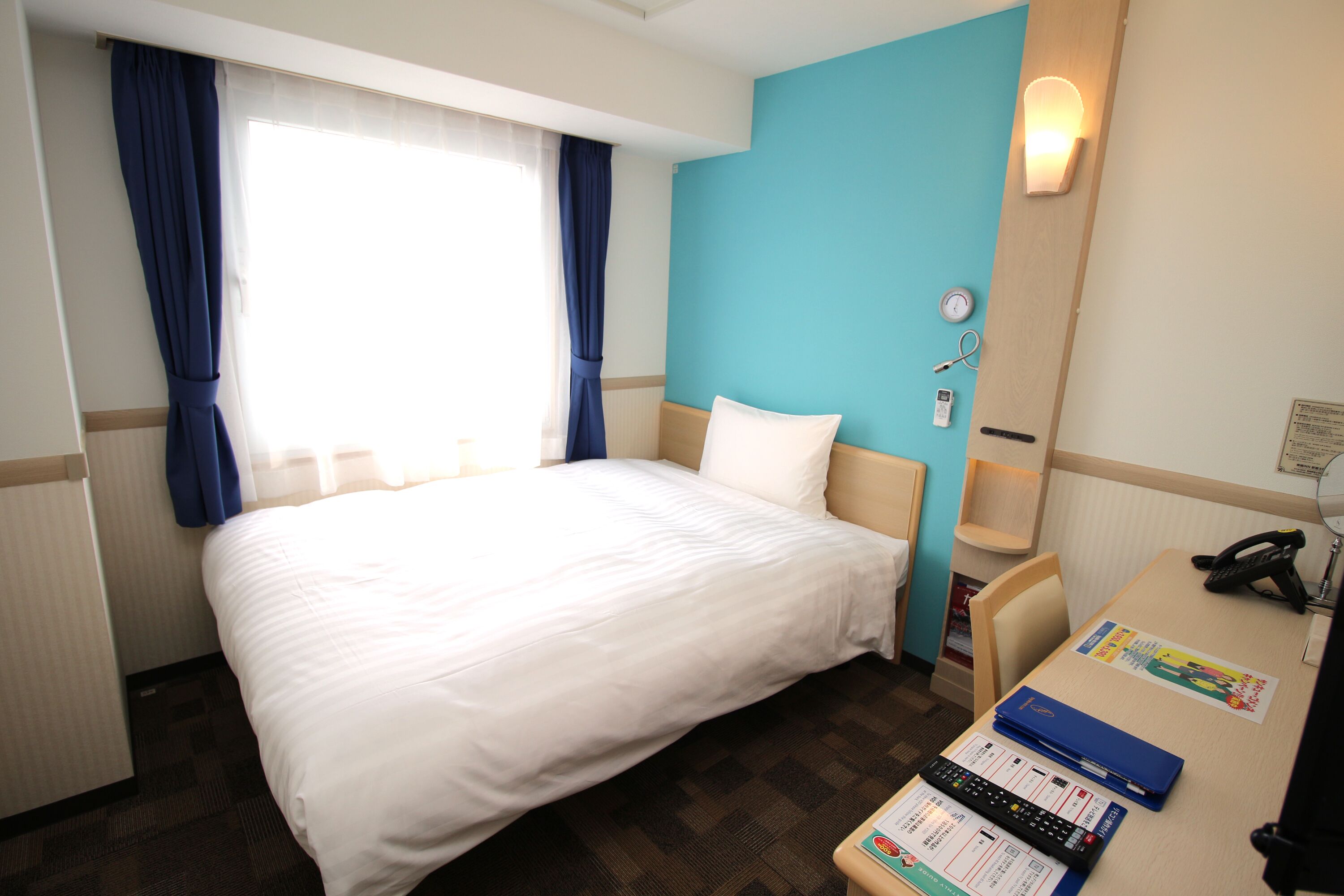 A hotel room with a large bed, a desk with a telephone and a computer, and a window with curtains.