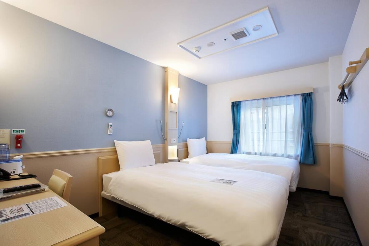 Ein Hotelzimmer mit zwei Betten, einem Schreibtisch mit Stuhl, einem kleinen Tisch, einem Fenster mit Vorhängen und einer Deckenleuchte.