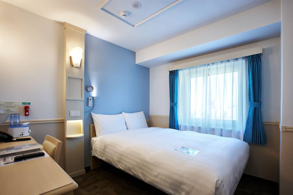 Ein Hotelzimmer mit einem Bett, einem Schreibtisch mit Telefon und Fernbedienung, einer Wandleuchte und einem Fenster mit blauen Vorhängen.