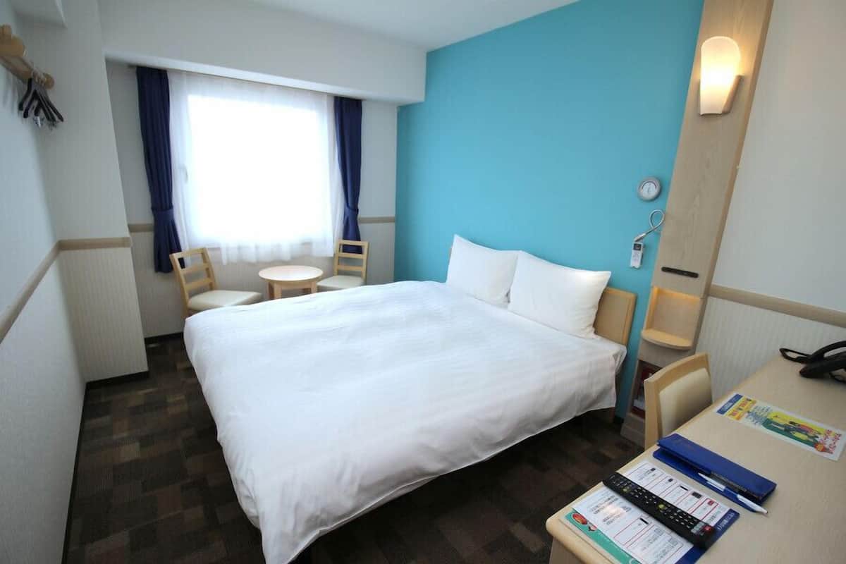 Ein Hotelzimmer mit einem großen Bett, einem Schreibtisch mit Stuhl, einem kleinen Tisch und einem Fenster mit Vorhängen.