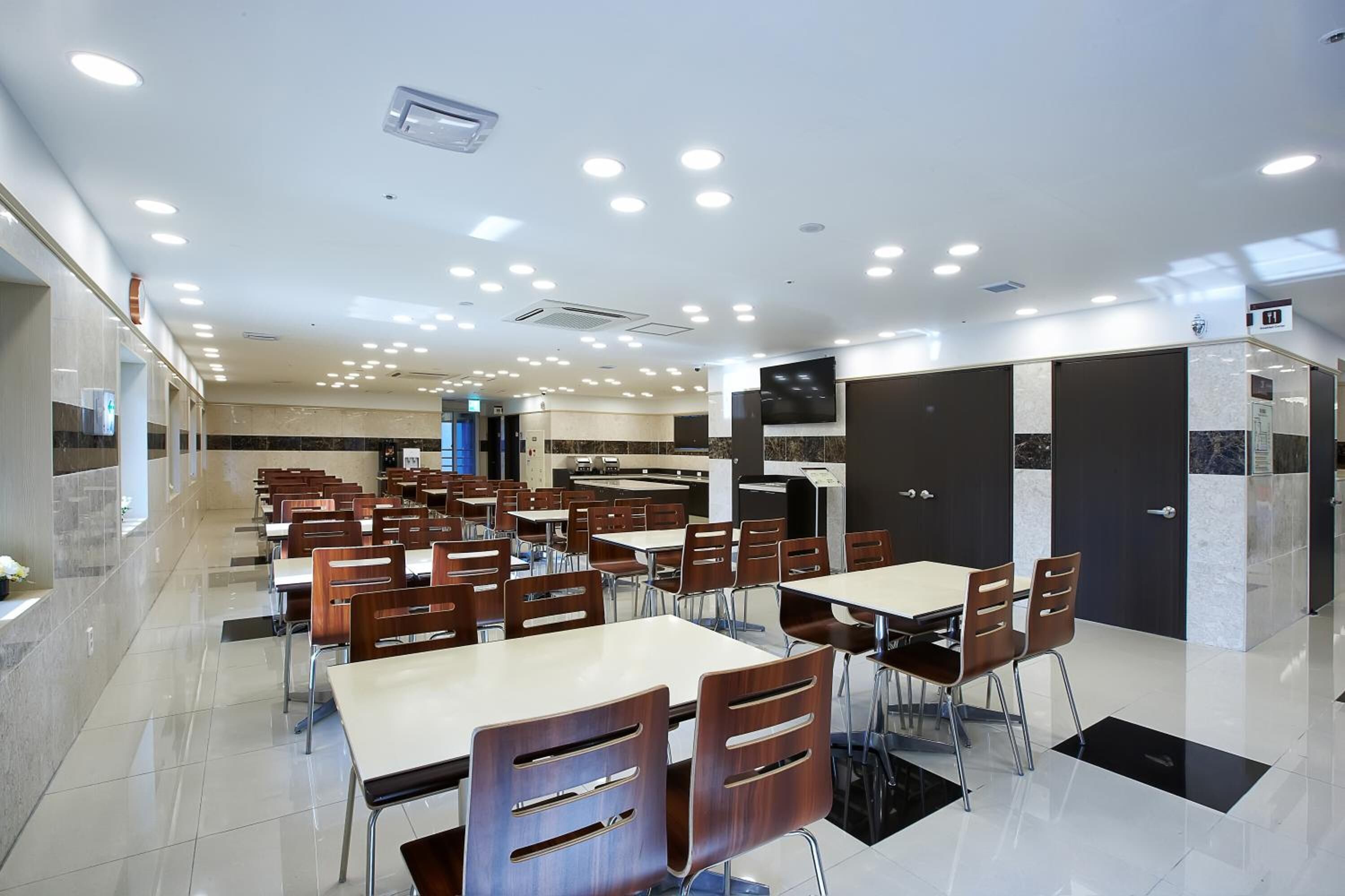 A modern dining area with multiple tables and chairs, a kitchen area, and a television screen on the wall.