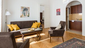 A living room with a sofa, armchairs, a coffee table, and a rug. There is a painting on the wall and a door leading to a kitchen. - KrakowRentals - Belle Epoque 1 Apartment (Kraków)