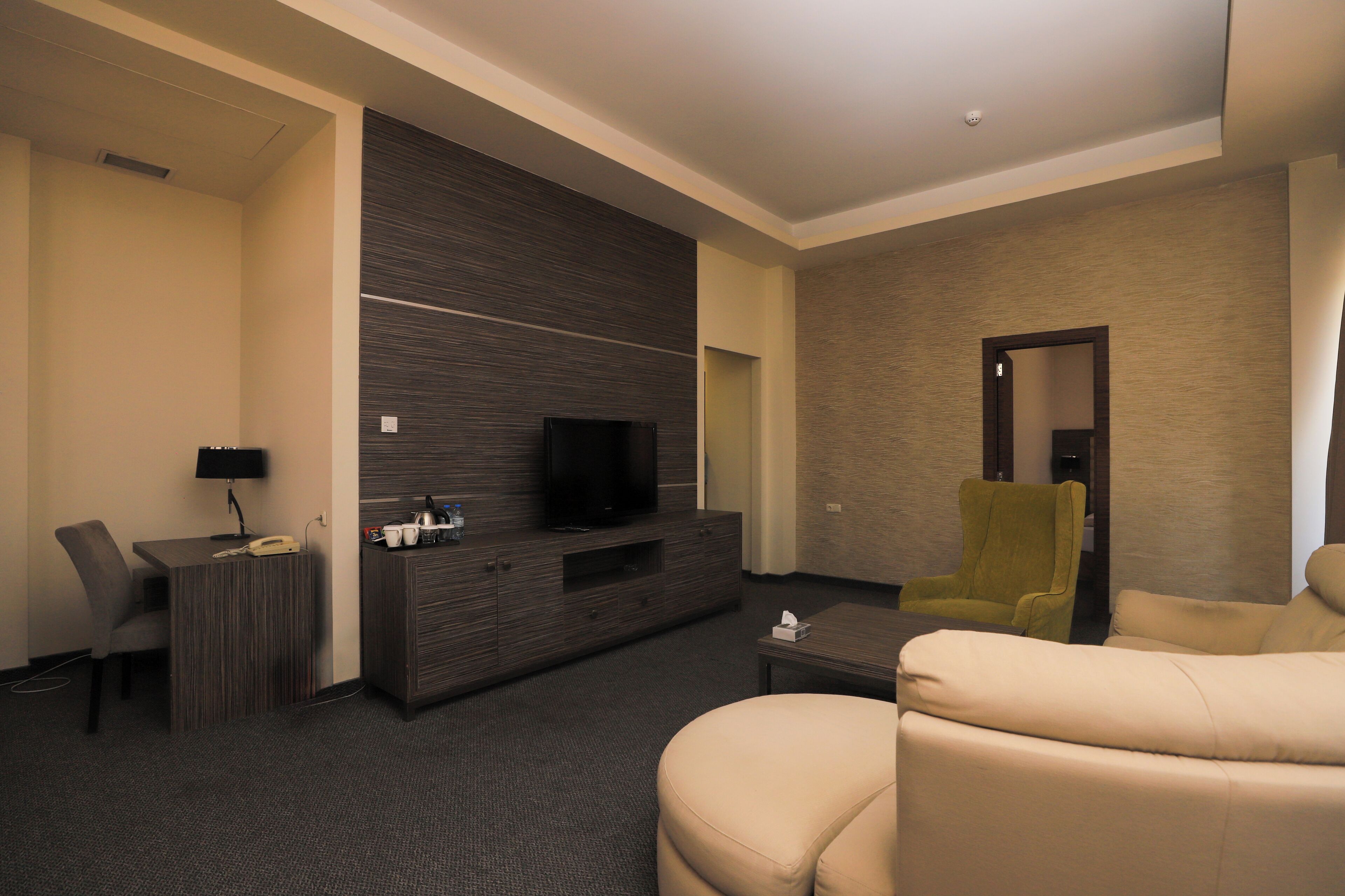 A modern hotel room with a flat-screen TV, a sofa, a chair, a small table, and a desk with a lamp.