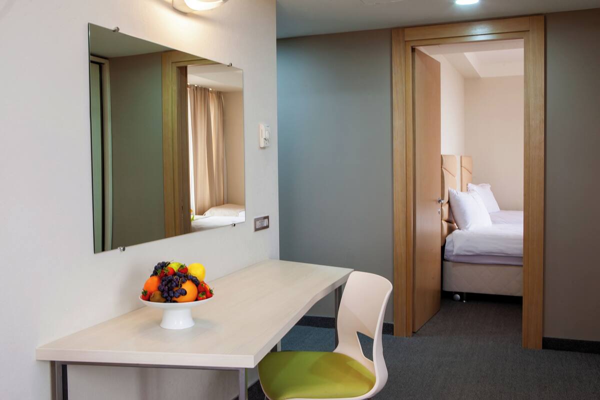 Une chambre d’hôtel avec un lit, une table de chevet sur laquelle se trouve un panier de fruits, une chaise et un miroir au mur.