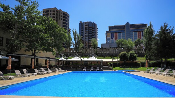 A swimming pool with lounge chairs and umbrellas, surrounded by trees and buildings.