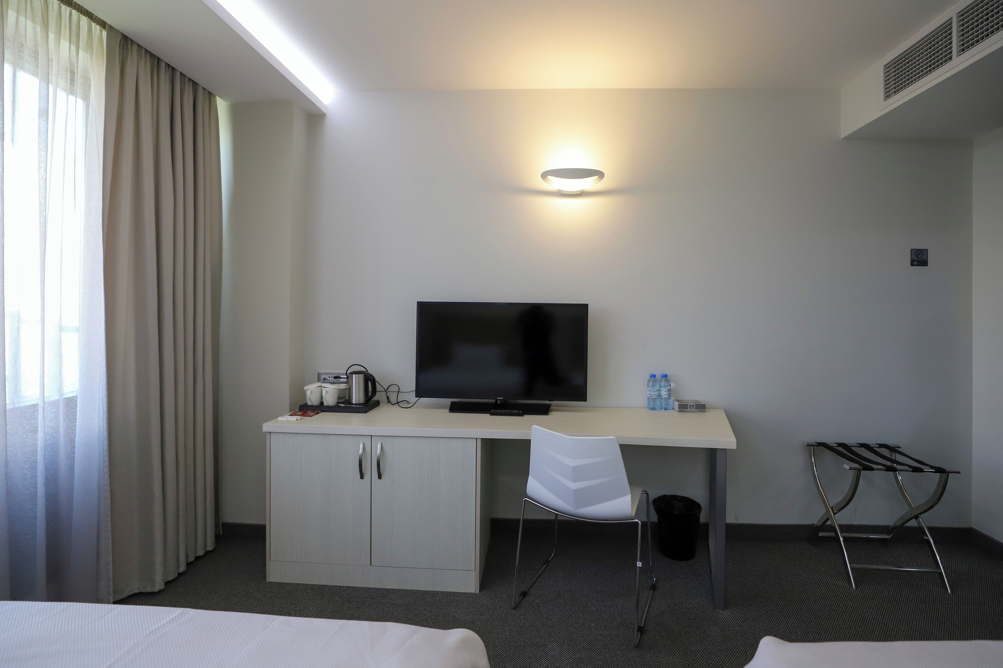 A hotel room with a bed, a desk with a television, a chair, a small cabinet, a wall-mounted light, and a window with curtains.