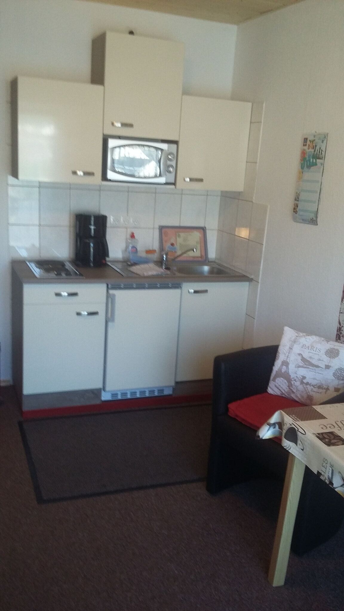 Une petite cuisine équipée d’un four à micro-ondes, d’une machine à café et d’un petit réfrigérateur. On y trouve une chaise avec un coussin rouge et une table sur laquelle est posée une publicité pour du café.