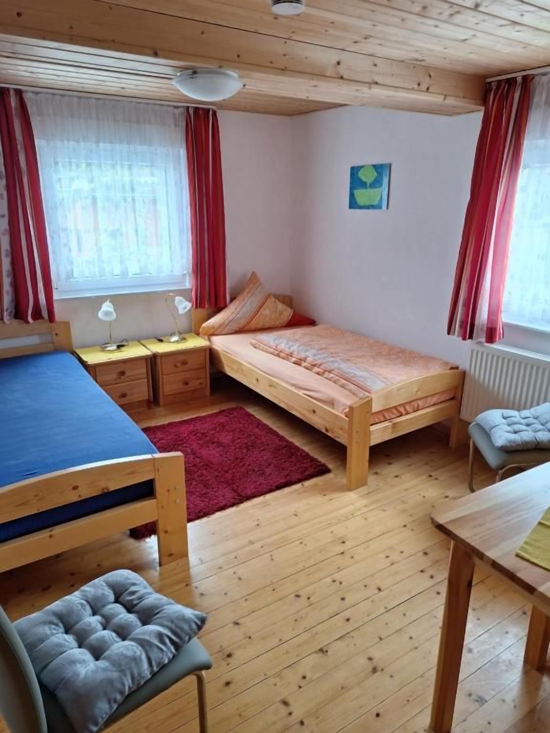 A room with two beds, a desk, and a chair. There are two windows with curtains, a radiator, and a wall-mounted picture.