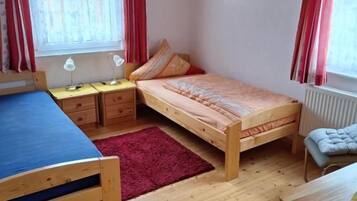 A room with two beds, a desk, and a chair. There are two windows with curtains, a radiator, and a wall-mounted picture.