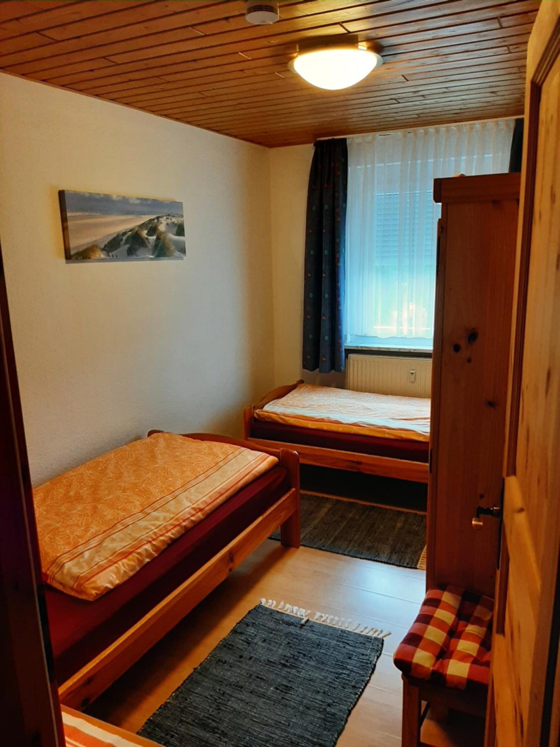 Une petite pièce chaleureuse, avec deux lits en bois, une fenêtre simple avec des rideaux, un radiateur et une armoire en bois.