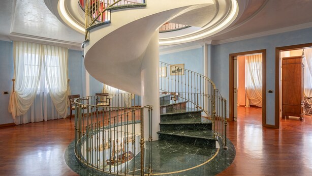 Una majestuosa escala espiraltra amb graons de marbre i barana de metall ornamentada, que condueix a un pis superior en una habitació àmplia amb terres de fusta i parets blaves clares.