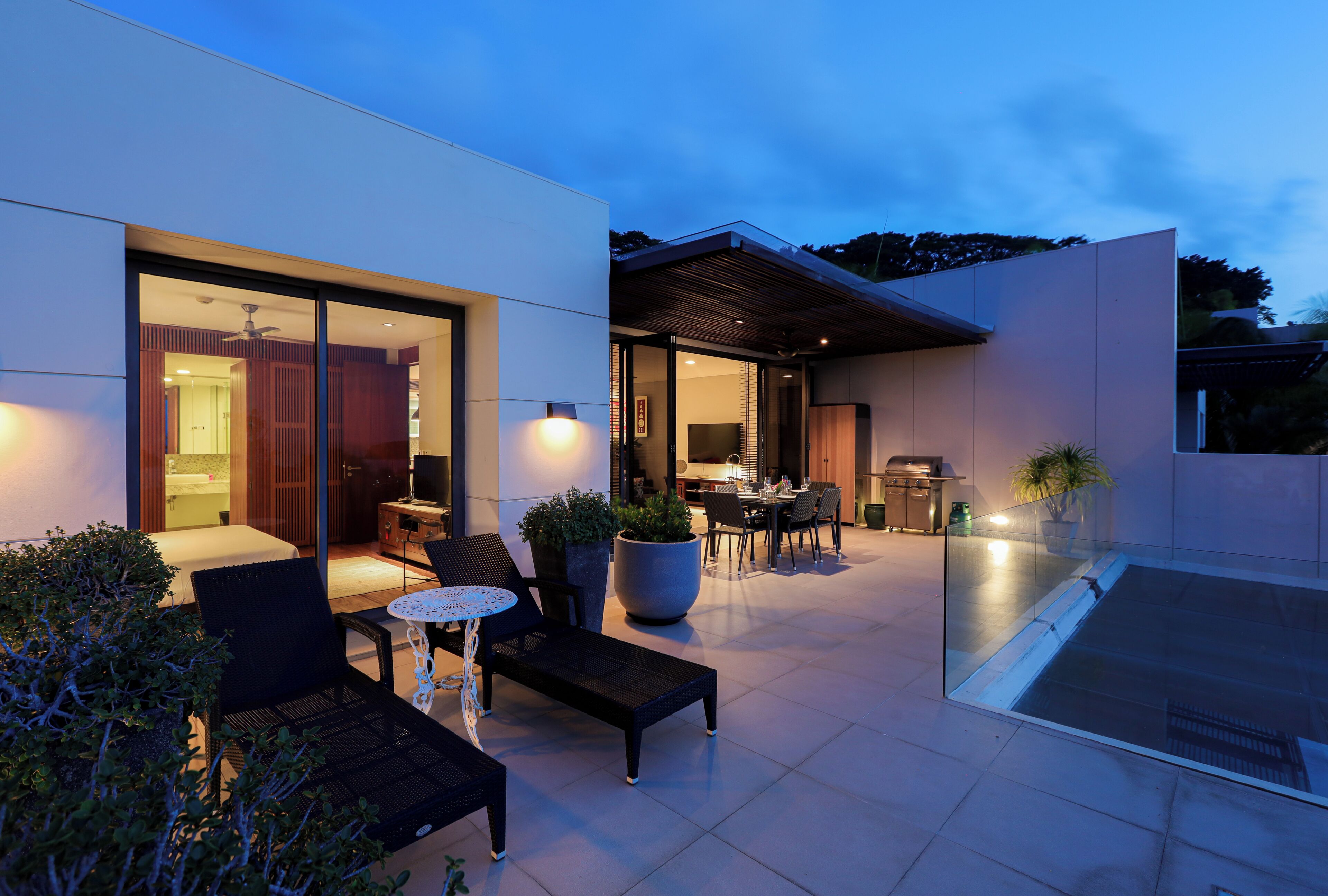 Um pátio externo moderno com área de estar, espaço para refeições e cozinha visível através das portas de vidro.