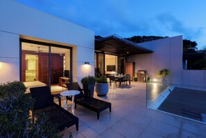 Um pátio externo moderno com área de estar, espaço para refeições e cozinha visível através das portas de vidro.