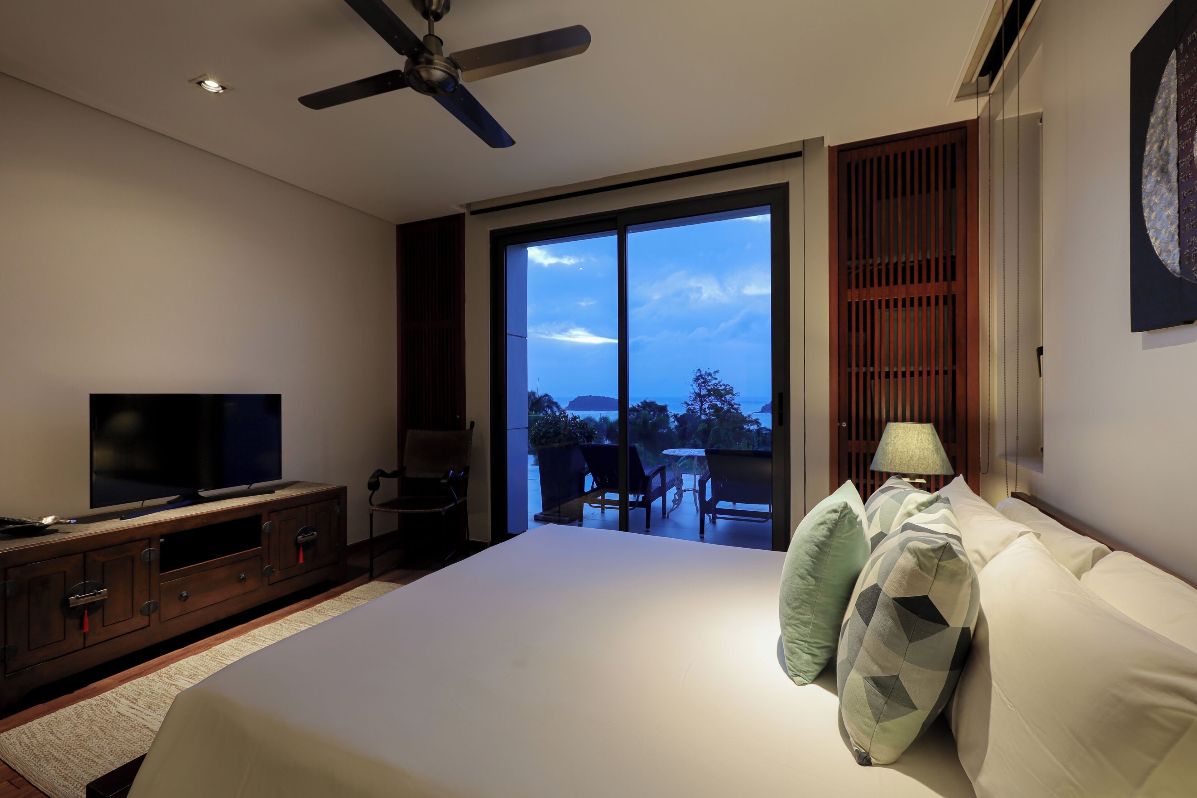 Quarto com cama, televisão, ventoinha no teto e vista para o exterior por uma porta de correr.