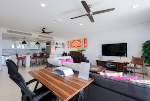 A modern living room with a wooden dining table, black chairs, a grey sofa, a flat-screen TV, ceiling fans, and a kitchen area in the background.