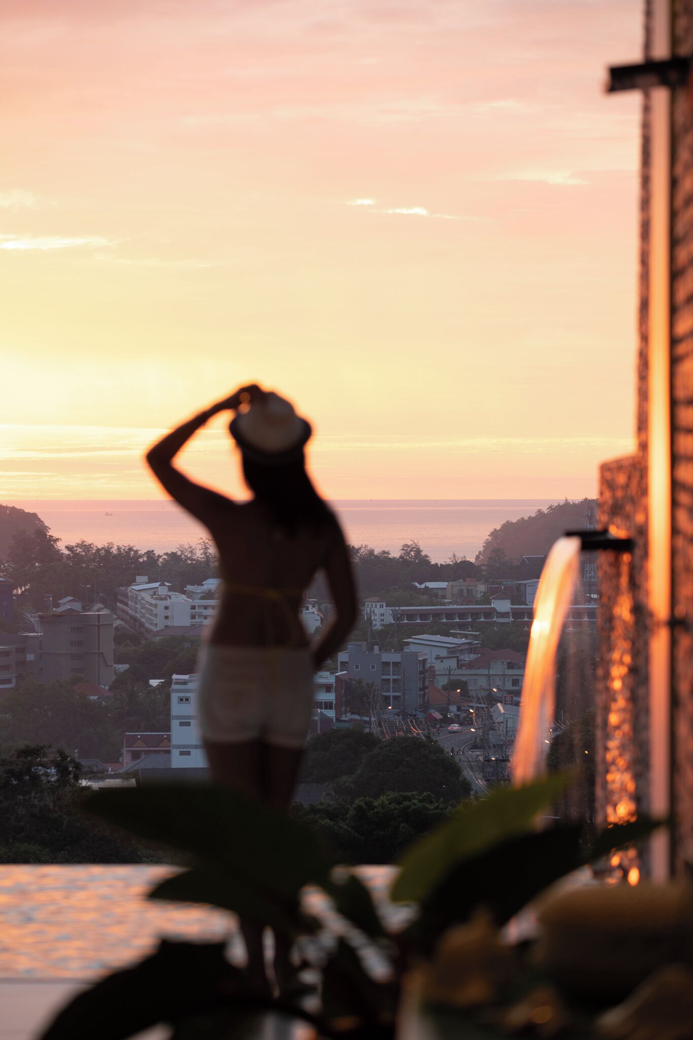 Silueta de una persona de pie en un balcón con vistas a un paisaje urbano y al océano al atardecer.