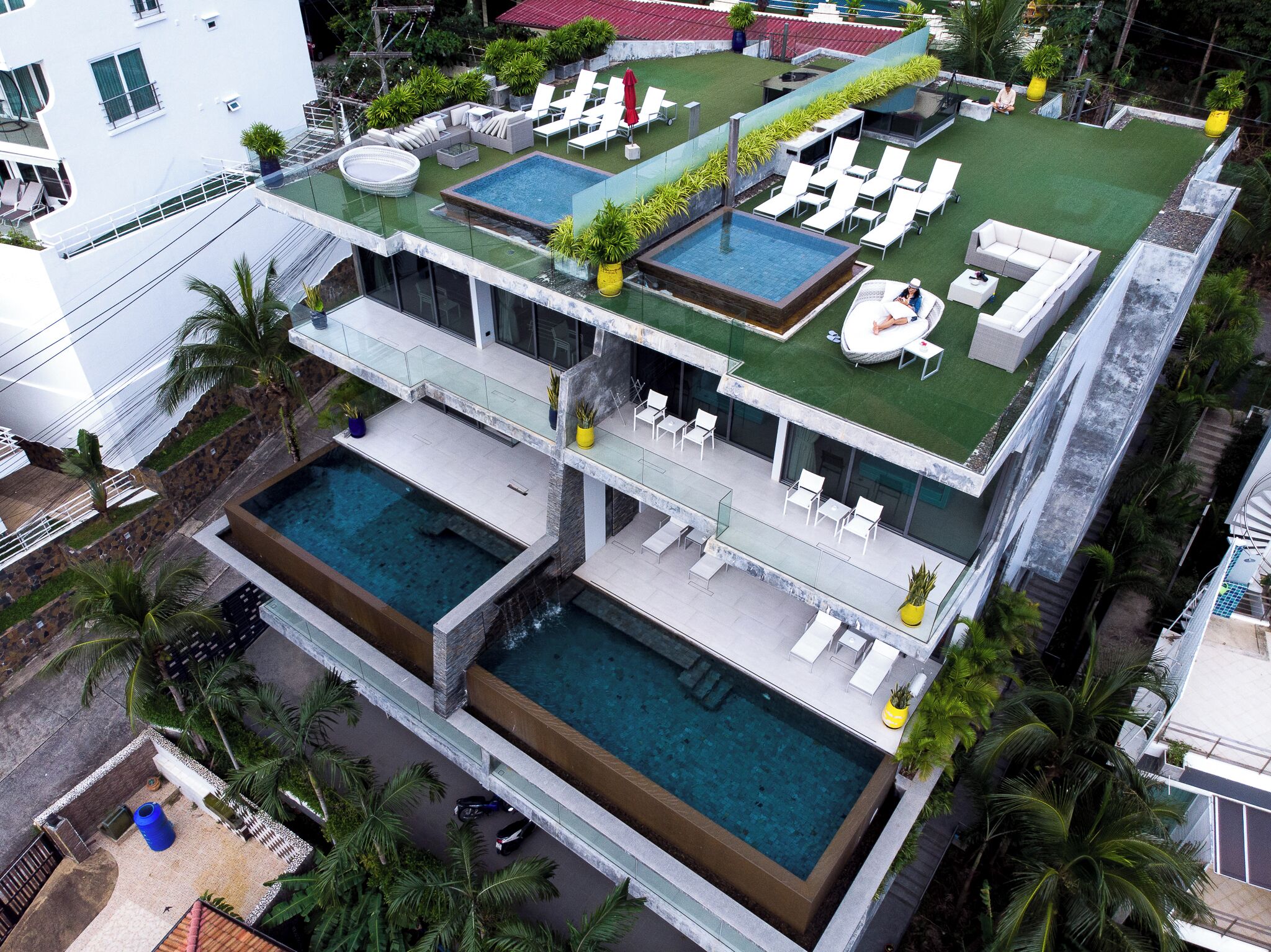 A modern multi-story hotel with two swimming pools, outdoor lounge areas, and a rooftop garden.