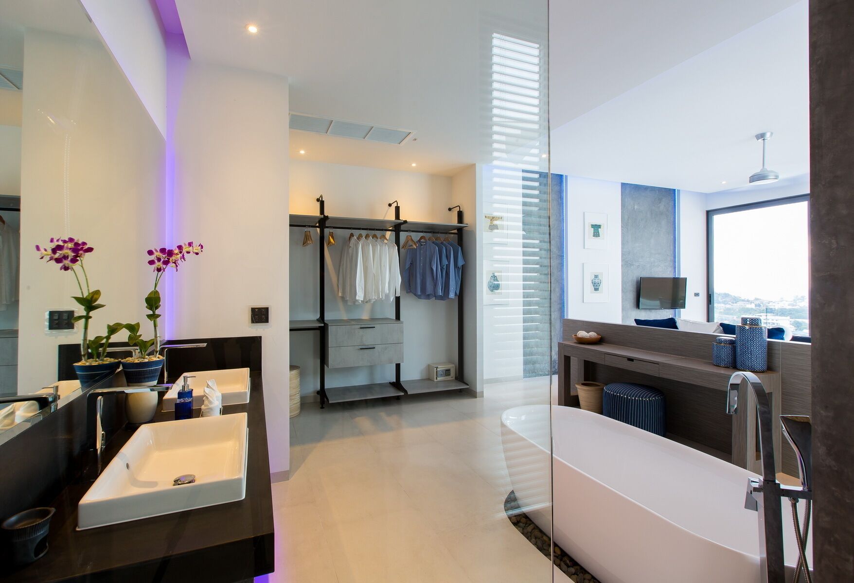 A modern bathroom with dual sinks, a vanity, a shower area, and a walk-in closet with hanging clothes.