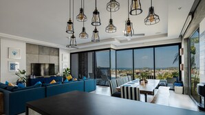 A modern living room with a large TV, blue sofas, a dining area, and large windows with a city view. - Villa Momo Private Pool near Kata Beach (Karon)