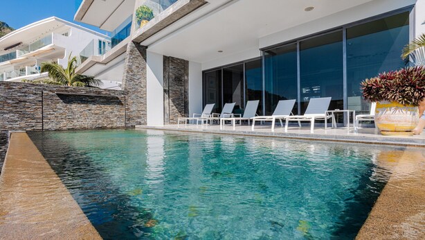 A modern pool area with clear water, white lounge chairs, and a stone wall. - Villa Momo Private Pool near Kata Beach (Karon)