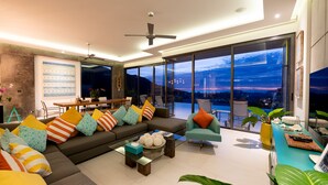 A modern living room with a large sofa, colorful cushions, a glass coffee table, and a view of the city at dusk. - Villa Omari Luxury Seaview Pool & Game Room (Karon)