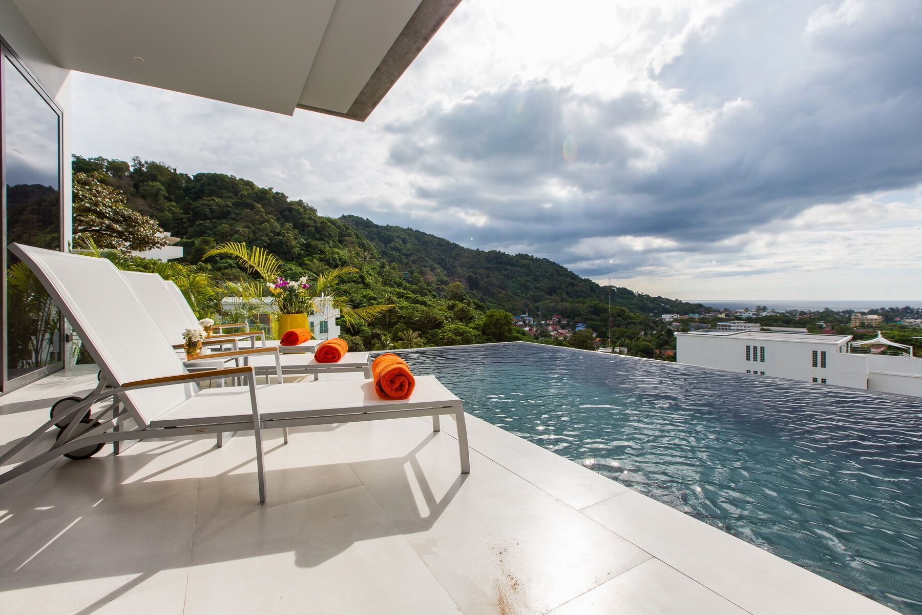 A rooftop infinity pool with white lounge chairs and orange towels, overlooking a lush hillside and a distant cityscape.