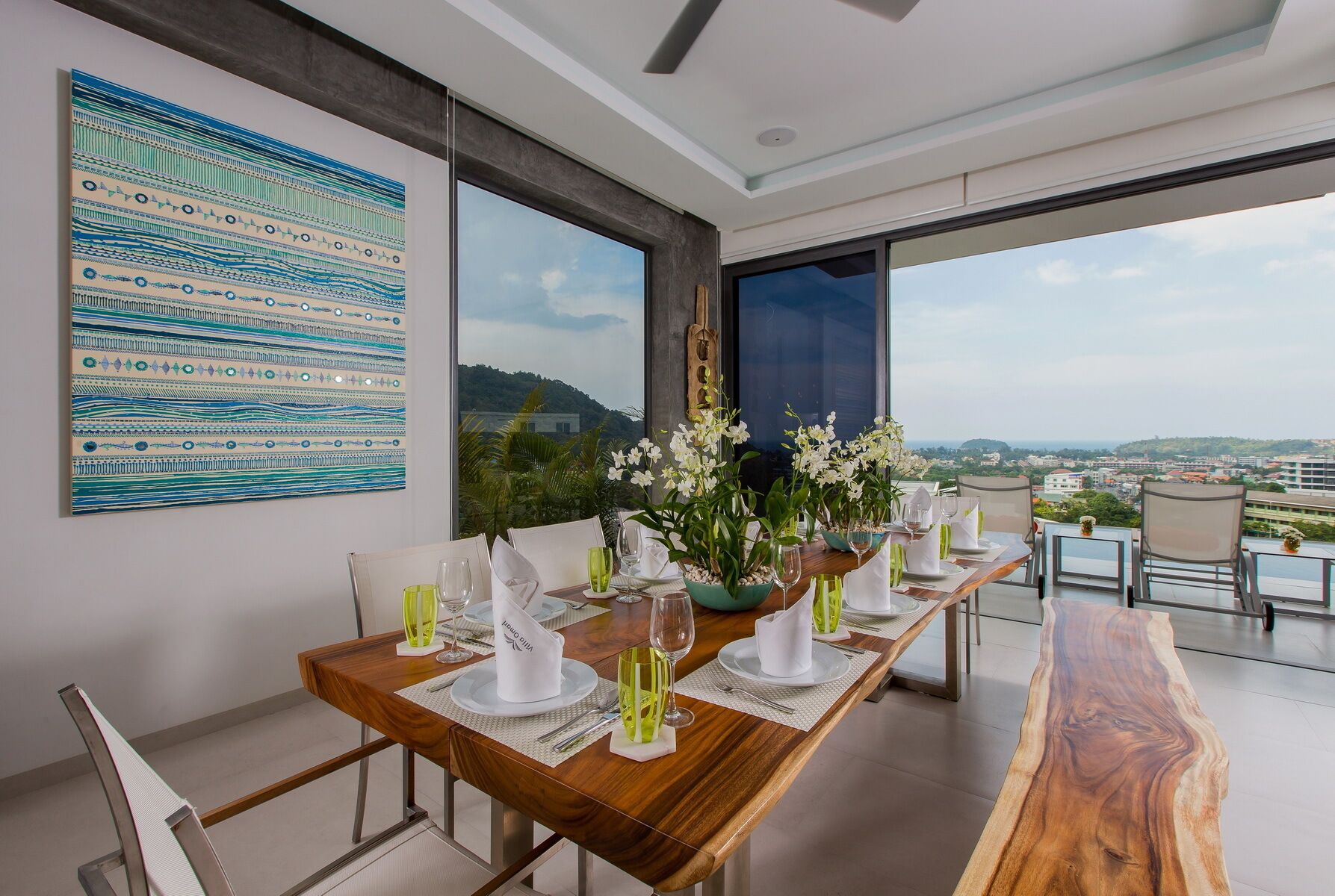 A dining area with a long wooden table set for six, large windows offering a city view, and a large abstract painting on the wall.