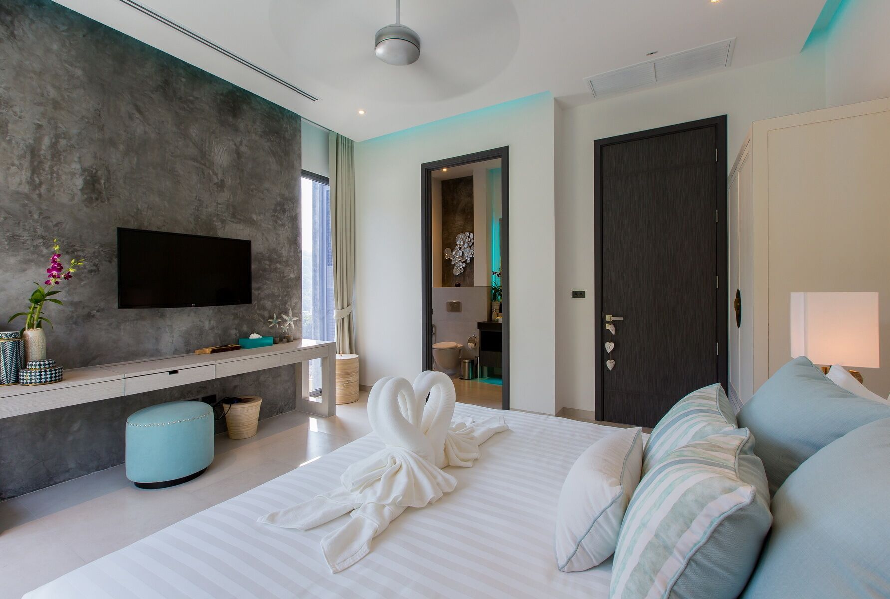 A modern hotel room with a large bed, a TV mounted on the wall, a desk with a vase of flowers, and a bathroom visible through an open door.