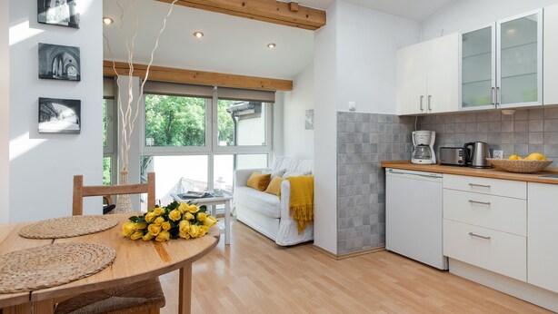 Una cucina moderna con mobili bianchi, un tavolo da pranzo in legno con sottopiatti intrecciati, e vista su un divano bianco e fiori gialli dalla finestra.
