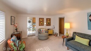 A living room with a grey sofa, a round ottoman, a small side table with a lamp, and a chair. There is a door leading to another room and a window with blinds. - 1 Br70's Inspired Comfy Condoclose to Broadmoor (Colorado Springs)