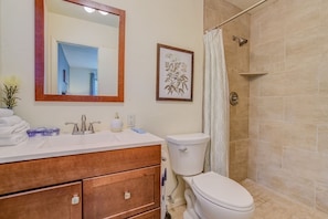 Une salle de bain avec un meuble-lavabo en bois, un lavabo blanc et des toilettes. On y trouve une douche avec des carreaux de couleur beige et une porte en verre.