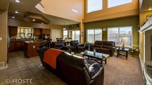 A spacious living area with a large leather sofa, a glass coffee table, and a dining area with chairs. There is a kitchen in the background with wooden cabinets and a large island. The room has large windows and a view of the outdoors. - 5BR Mtn Views Family Getaway HOT TUB (Colorado Springs)