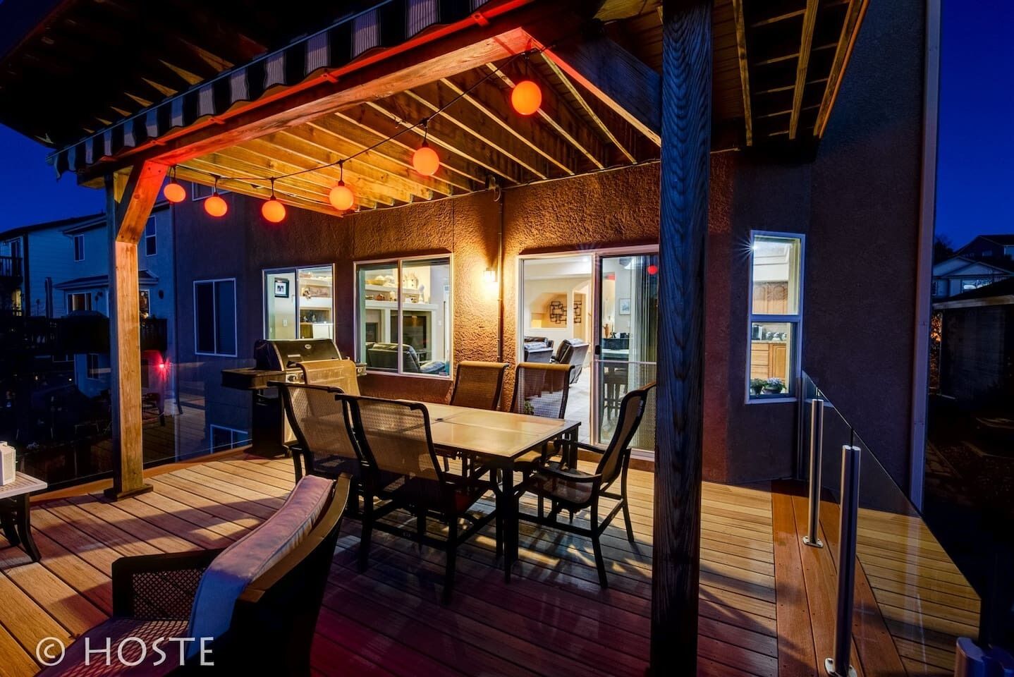 An outdoor patio with a wooden table and chairs, a grill, and hanging orange lights.
