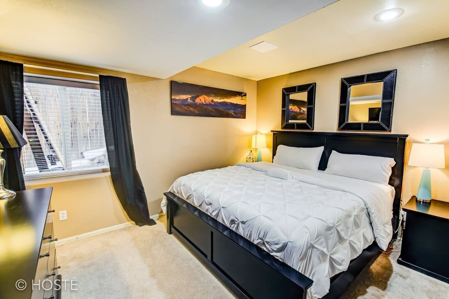 Une chambre à coucher avec un lit, une table de chevet, une lampe, une fenêtre avec des rideaux et un tableau au mur.