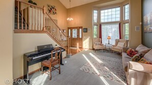 A spacious living room with a grand piano, a staircase, and large windows allowing natural light.