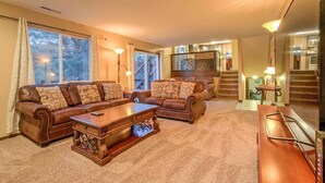 A spacious living room with a large brown leather sofa set, a wooden coffee table, and a television. There is a staircase leading to an upper level, and a dining area is visible in the background. - 4BR Entertainer AF Academy Hot Tub, Firepit! (Colorado Springs)