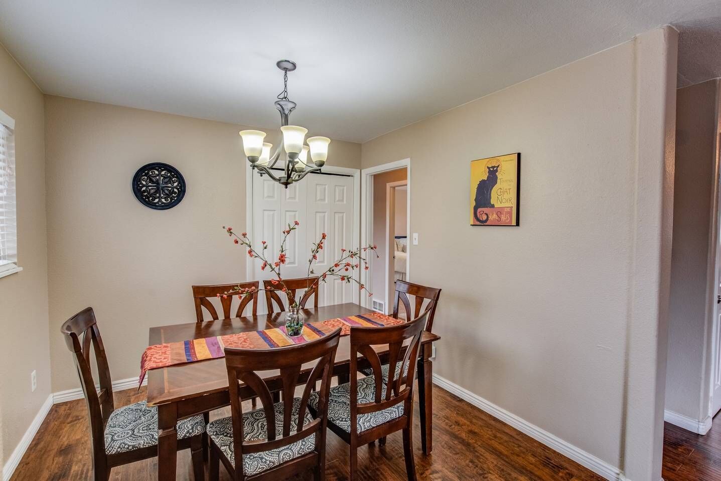 A dining room with a wooden table, six chairs, a chandelier, a vase with flowers, a wall clock, and a framed picture.