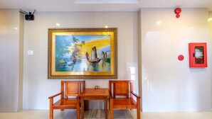 A room with a large framed painting of a tropical scene, a wooden table with two chairs, and a red fire alarm on the wall. - Dubai Hotel (Da Nang)